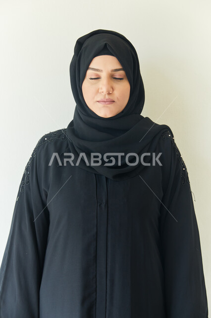التأمل والتفكير وإسترجاع الذكريات، بورتريه مقرب لشابه عربيه خليجيه اماراتيه محجبه ترتدي عباءه سوداء تقف بإستقامه وتغمض عينيها، ايماءات وجه تدل على الشعور بالنعاس، خلفيه بيضاء.