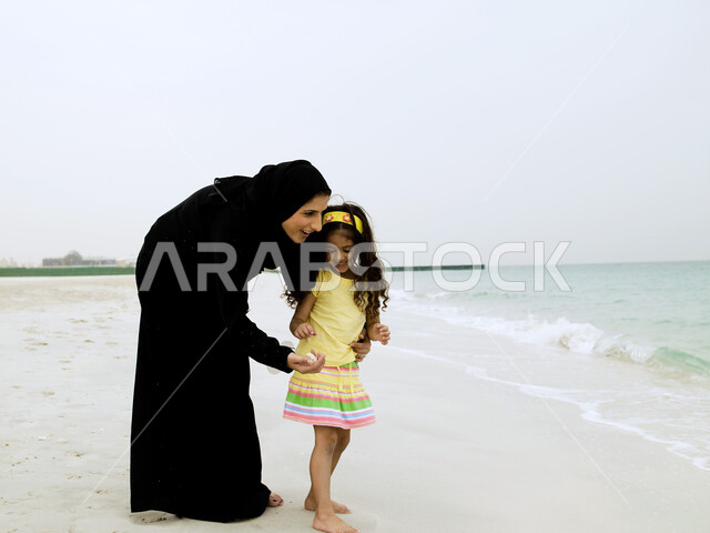صورة الجسم بالطول الكامل, قضاء أجواء أسرية ممتعة, المشي على رمال شاطئ البحر, تغيير الاجواء المنزلية وزيادة الترابط الاسري بين الابناء والاهل، السياحة في دول الخليج العربي, امرأة عربية خليجية إماراتية محجبة تقضي وقت ممتع مع إبنتها على ساحل البحر