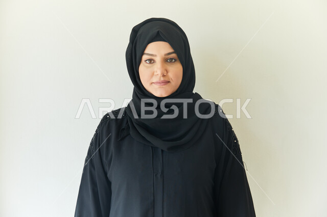 أخذ صوره للوثائق الرسميه، ايماءات وجه تدل على السعاده، الإهتمام بالمظهر الخارجي، بورتريه مقرب لشابه عربيه خليجيه اماراتيه محجبه ترتدي عباءه سوداء تقف بإستقامه أمام الكاميرا، خلفيه بيضاء.