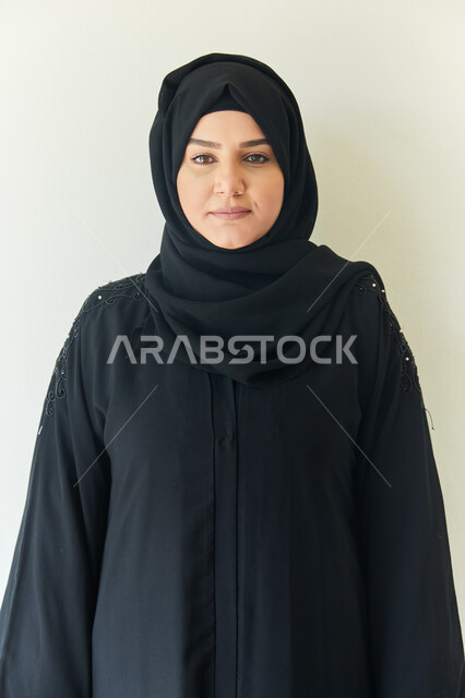 ايماءات وجه تدل على السعاده، الإهتمام بالمظهر الخارجي، إلتقاط صوره للوثائق الرسميه، بورتريه مقرب لشابه عربيه خليجيه اماراتيه محجبه ترتدي عباءه سوداء تقف بإستقامه أمام الكاميرا، خلفيه بيضاء.