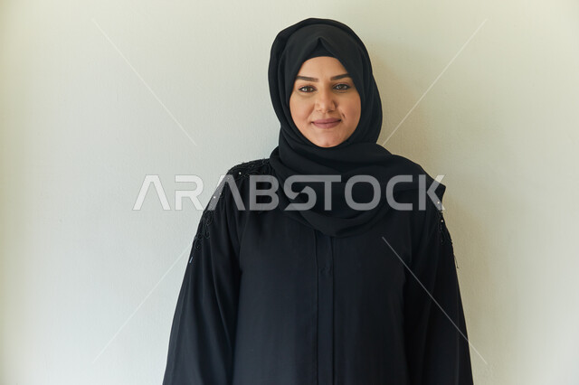 إلتقاط صوره للوثائق الرسميه، بورتريه مقرب لشابه عربيه خليجيه اماراتيه محجبه ترتدي عباءه سوداء تقف بإستقامه أمام الكاميرا، الإهتمام بالمظهر الخارجي، ايماءات وجه تدل على السعاده، خلفيه بيضاء.