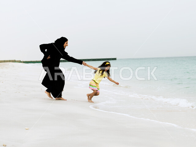 قضاء وقت ممتع مع الابناء, مفهوم الاسرة السعودية السعيدة المترابطة، السياحة في دول الخليج العربي والاستمتاع بالاجازة الصيفية، صورة الجسم بالطول الكامل, إمرأة عربية خليجية اماراتية محجبة ترتدي العباءة السوداء تقضي وقت ممتع مع ابنتها على الشاطئ