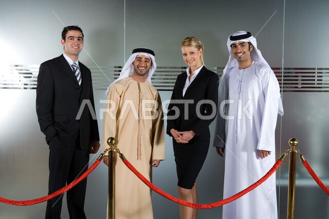 حاجز الحبل الأحمر المعلق بدعامات فولاذية، رجلان أعمال عربيان خليجيان إماراتيان يرتديان الزي الإماراتي التقليدي ويقفان مع رجل وسيدة أعمال اجنبيان لأخذ صورة تذكارية، تحقيق إستراتيجيات وأهداف العمل، مفهوم التعاون والشراكة بين الشركات الإماراتية والأجنبية
