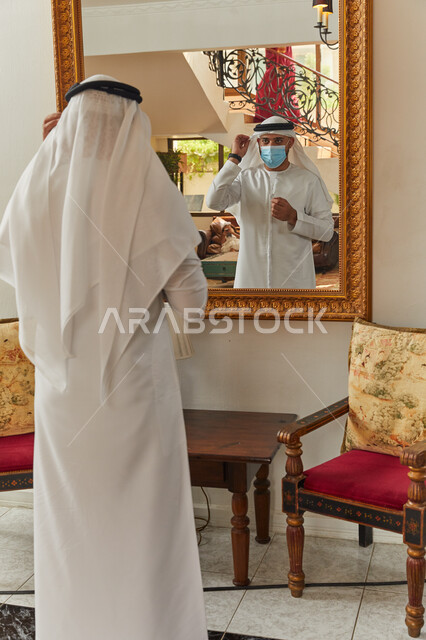 الاعتناء والاهتمام بالمظهر الخارجي، فلل وشقق سكنية فاخرة في الامارات، رجل عربي خليجي اماراتي يرتدي الكندورة والغترة يقف على المرآة يقوم بترتيب ثيابه، الاستعداد للخروج من المنزل، الالتزام بالاجراءات الاحترازية وارتداء الكمامة للوقاية من فايروس كورونا.