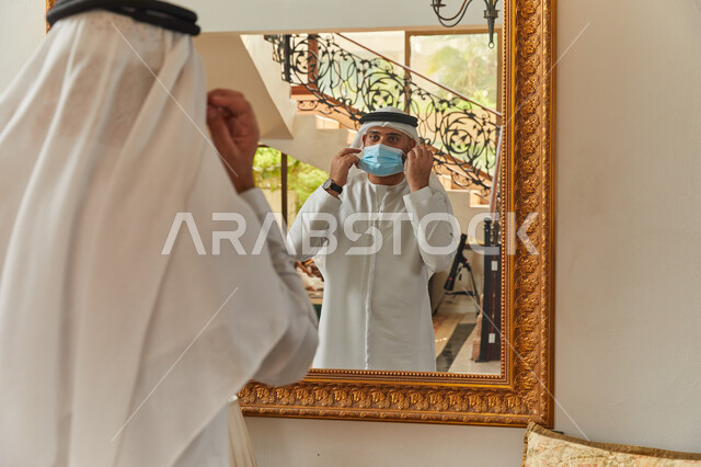 الاستعداد للخروج من المنزل، ترتيب المظهر الخارجي من خلال انعكاس المرآة، فلل وشقق سكنية فاخرة في الامارات، رجل عربي خليجي اماراتي يرتدي الكندورة والغترة يقوم بارتداء الكمامة، الالتزام بالاجراءات الاحترازية  للوقاية من الامراض وفايروس كورونا.