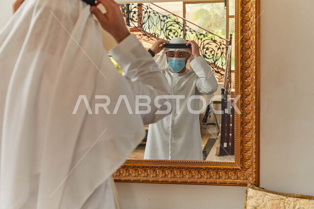 Paying attention and taking care of the outward appearance, luxury villas and apartments in the UAE, an Emirati Arab man wearing a kandora and a ghutra standing at the mirror arranging his clothes, getting ready to leave the house, adhering to the precaut