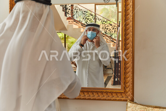التجهز والاستعداد للخروج من المنزل، ترتيب المظهر الخارجي من خلال انعكاس المرآة، فلل وشقق سكنية فاخرة في الامارات، رجل عربي خليجي اماراتي يرتدي الكندورة والغترة يقوم بارتداء الكمامة، الالتزام بالاجراءات الاحترازية  للوقاية من الامراض وفايروس كورونا.