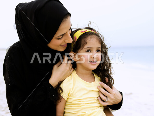 مشاعر الحب و العطف, قضاء أوقات عائلية ممتعة بالعطلة الصيفية على شاطئ البحر, صورة مقربة لأم عربية خليجية إماراتية محجبة ترتدي العباءة السوداء  تحمل وتنظر إلى إبنتها بحب وحنان، مفهوم الأمومة والطفولة