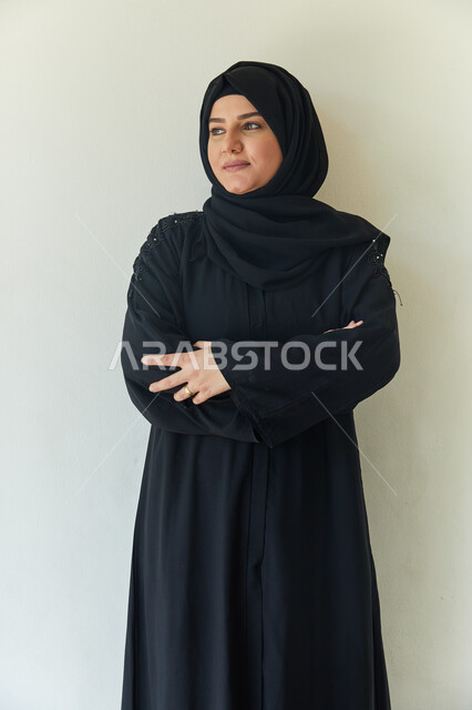 الإهتمام والعنايه بالمظهر الخارجي، ايماءات وجه تدل على السعاده والسرور، بورتريه مقرب لامرأه عربيه خليجيه اماراتيه محجبه ترتدي عباءه سوداء تبتسم وتقف بإستقامه أمام الكاميرا مكتفه اليدين، خلفيه بيضاء.