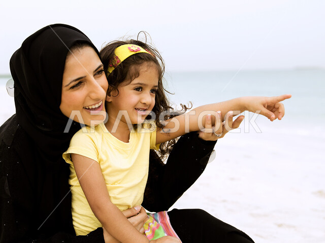 الاشارة والنظر إلى شيء ما, قضاء أوقات عائلية ممتعة بالعطلة الصيفية على شاطئ البحر, صورة مقربة لأم عربية خليجية إماراتية محجبة تحمل إبنتها بحب وحنان، مفهوم الأمومة والطفولة, مشاعر الحب و العطف