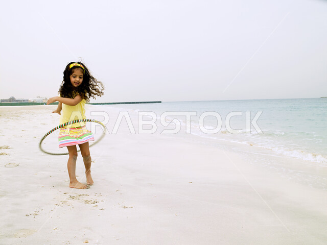 قضاء يوم العطلة على البحر, تعزيز المهارات الحركية للاطفال عن طريق اللعب, الاستمتاع بالسباحة على شاطئ البحر, فتاة صغيرة عربية خليجية إماراتية تلعب بطوق الهولاهوب على الشاطئ, ارتداء الملابس الصيفية