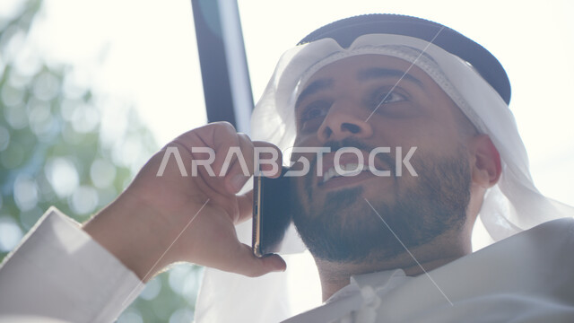مكالمات ودردشات عبر مواقع التواصل الاجتماعي, صورة مقربة لرجل عربي خليجي إماراتي مبتسم يرتدي الكندورة والغترة يقوم بإجراء مكالمة عبر الهاتف المحمول, التواصل مع الأصدقاء عبر الانترنت بإيماءات الفرح والسرور