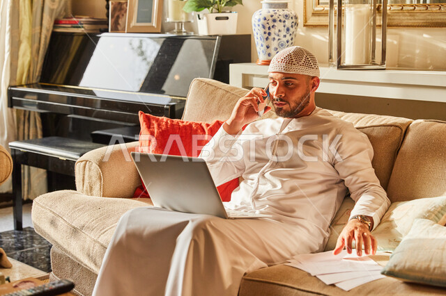 متابعة الاعمال والمهام من المنزل في ظل جائحة كورونا، الاجابة على الاستفسارات عبر الجوال، رجل عربي خليجي اماراتي يرتدي الكندورة و الطاقية الاماراتية البيضاء يجلس على الاريكة في بيته يقوم بإنجاز العمل اونلاين من خلال الحاسب المحمول.