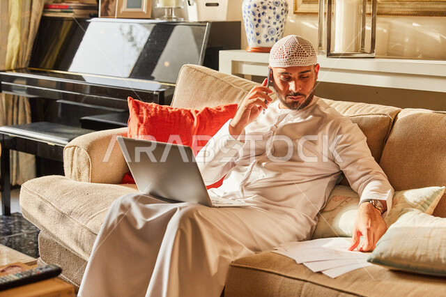 الاجابة على الاستفسارات عبر الجوال، إنجاز العمل اونلاين من خلال الحاسب المحمول، رجل عربي خليجي اماراتي يرتدي الكندورة و الطاقية الاماراتية البيضاء يجلس على الاريكة في بيته يقوم بالبحث بين اوراق البيانات، اتمام الاعمال والمهام من المنزل في ظل جائحة كورونا.