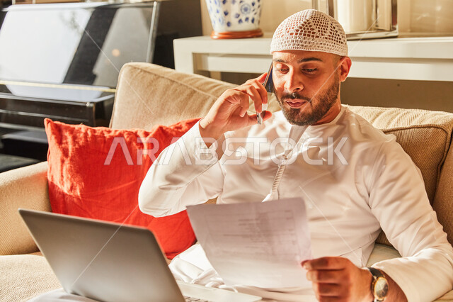  إنجاز العمل اونلاين من خلال الحاسب المحمول، رجل عربي خليجي اماراتي يرتدي الكندورة و الطاقية الاماراتية البيضاء يجلس على الاريكة في بيته يقوم بقراءة المعلومات من الورقة، الرد على الاستفسارات عبر الجوال، اتمام الاعمال والمهام من المنزل في ظل جائحة كورونا.