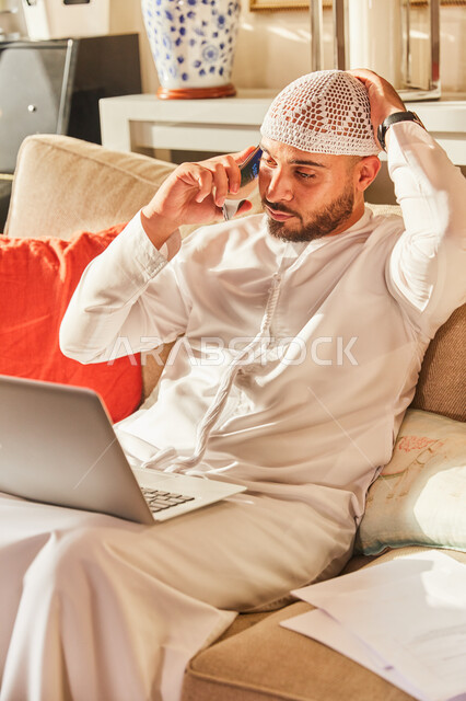 تلقي اخبار صادمة ومفاجئة، الرد على الاستفسارات عبر الجوال، اتمام الاعمال والمهام من المنزل في ظل جائحة كورونا، رجل عربي خليجي اماراتي يرتدي الكندورة و الطاقية الاماراتية البيضاء يجلس على الاريكة في بيته يقوم بإنجاز العمل اونلاين من خلال الحاسب المحمول.