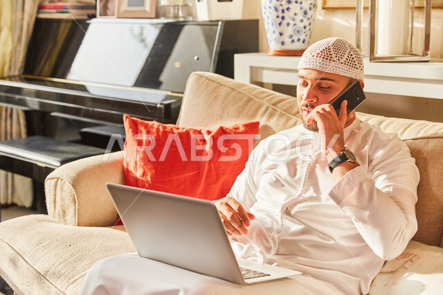  الرد والاجابة على الاستفسارات عبر الجوال، اتمام الاعمال والمهام من المنزل في ظل جائحة كورونا، رجل عربي خليجي اماراتي يرتدي الكندورة و الطاقية الاماراتية البيضاء يجلس على الاريكة في بيته يقوم بإنجاز العمل اونلاين من خلال الحاسب المحمول.