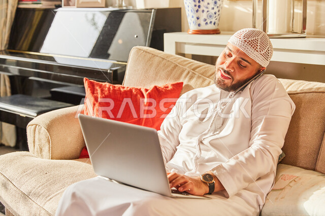 اتمام الاعمال والمهام من المنزل في ظل جائحة كورونا، رجل عربي خليجي اماراتي يرتدي الكندورة و الطاقية الاماراتية البيضاء يجلس على الاريكة في بيته ويقوم بالعمل من خلال الحاسب المحمول، الرد والاجابة على الاستفسارات عبر الجوال، تلقي اخبار هامة وسعيدة.