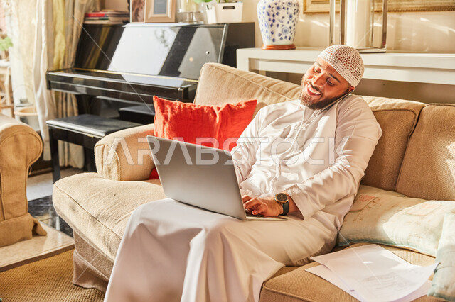 انهاء الاعمال والمهام من المنزل في ظل جائحة كورونا، رجل عربي خليجي اماراتي يرتدي الكندورة و الطاقية الاماراتية البيضاء يجلس على الاريكة في بيته ويقوم بالعمل من خلال الحاسب المحمول، الرد والاجابة على الاستفسارات عبر الجوال، تلقي اخبار هامة وسعيدة.