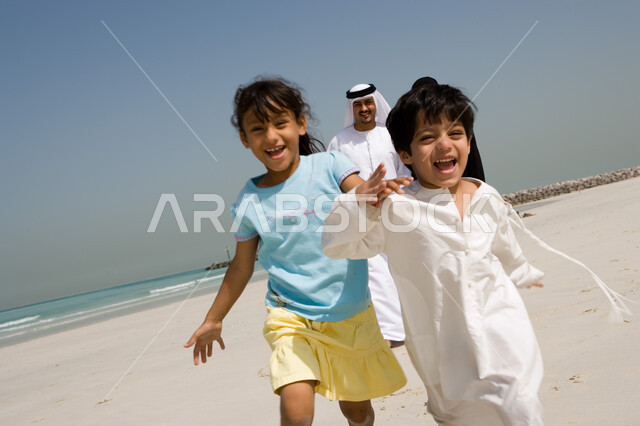 أخ وأخت إماراتيان يلعبان ويركضان على شاطئ البحر، أسرة عربية خليجية إماراتية تستمتع بالعطلة الصيفية، الأجواء العائلية المليئة بالحب، إيماءات السعادة والفرح بقضاء الوقت مع العائلة، فعاليات وأنشطة ترفيهية مسلية للأبناء، مفهوم الترابط الأسري