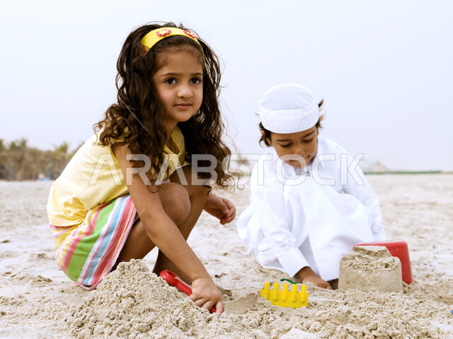  فعاليات ترفيهية مسلية للأطفال, أخ وأخت يلعبان على الشاطئ,قضاء يوم العطلة على البحر, فتاة بإيماءات وجه تدل على السعادة في اللعب برمال البحر الناعمة، طفل عربي خليجي إماراتي يرتدي الثوب السعودي التقليدي يقوم في بناء المجسمات الرملية
