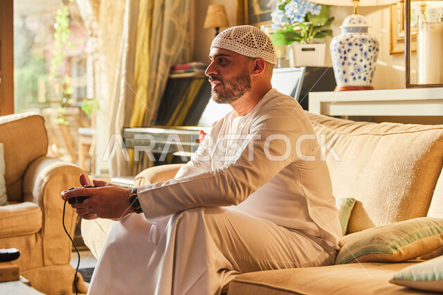 تقنيات وأجهزة الفيديو المسلية الحديثة، ايماءات الاندماج و التركيز، رجل عربي خليجي اماراتي يرتدي الكندورة و الطاقية البيضاء الاماراتية يجلس على الاريكة يمسك عصا التحكم بيده يقوم باللعب في ألعاب البلايستيشن الترفيهية، قضاء اوقات ممتعة في المنزل.