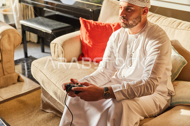 ايماءات الاندماج و التركيز، رجل عربي خليجي اماراتي يرتدي الكندورة و الطاقية البيضاء الاماراتية يجلس على الاريكة يمسك عصا التحكم بيده يقوم باللعب في ألعاب البلايستيشن الترفيهية، تقنيات وأجهزة الفيديو المسلية الحديثة، قضاء اوقات ممتعة في المنزل.