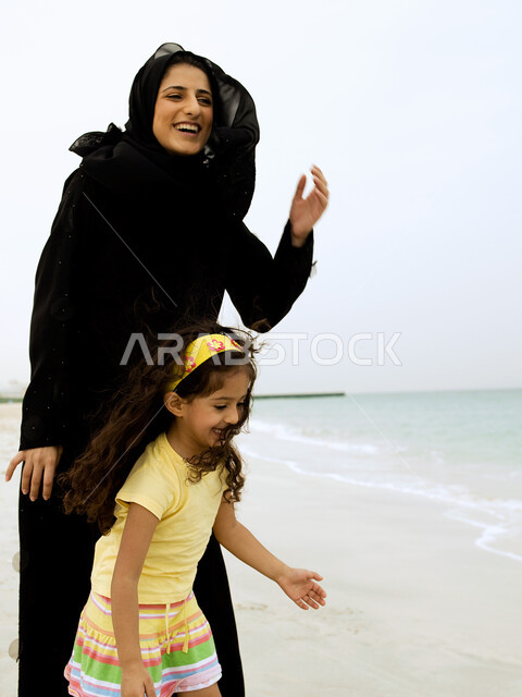 أم تلعب مع إبنتها بإيماءات وجه ويدين مختلفة تدل على السعادة، مفهوم الأمومة والطفولة، قضاء أوقات عائلية ممتعة بالعطلة الصيفية على شاطئ البحر, امرأة عربية خليجية إماراتية محجبة تقضي وقت ممتع مع إبنتها على ساحل البحر
