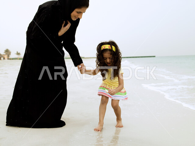 قضاء أجواء أسرية ممتعة, المشي على رمال شاطئ البحر, تغيير الاجواء المنزلية وزيادة الترابط الاسري بين الابناء والاهل، السياحة في دول الخليج العربي, امرأة عربية خليجية إماراتية محجبة تقضي وقت ممتع مع إبنتها على ساحل البحر