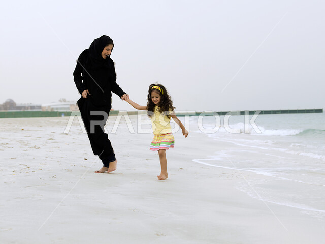 صورة الجسم بالطول الكامل, الاستجمام والاستمتاع بنسمات الهواء المختلفة على شواطئ البحر, الحصول على الاسترخاء والراحة عن طريق المشي على الرمال الناعمة على الساحل, امرأة عربية خليجية إماراتية محجبة تقضي وقت ممتع مع إبنتها على شاطئ البحر