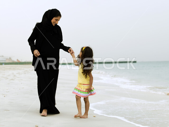 الحصول على الاسترخاء والراحة عن طريق المشي على الرمال الناعمة على الساحل, صورة الجسم بالطول الكامل, الاستجمام والاستمتاع بنسمات الهواء المختلفة على شواطئ البحر, امرأة عربية خليجية إماراتية محجبة تقضي وقت ممتع مع إبنتها على شاطئ البحر
