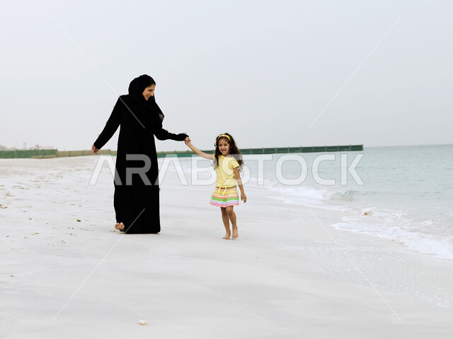 الاستجمام والاستمتاع بنسمات الهواء المختلفة على شواطئ البحر, الحصول على الاسترخاء والراحة عن طريق المشي على الرمال الناعمة على الساحل, صورة الجسم بالطول الكامل, امرأة عربية خليجية إماراتية محجبة تقضي وقت ممتع مع إبنتها على شاطئ البحر