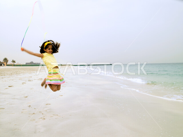 Spending fun times on vacation at the sea, enhancing children's motor skills by playing, enjoying swimming on the seashore, a close-up of a young Arab Emirati girl playing with a hula hoop and jumping high with enthusiasm on the sea coast