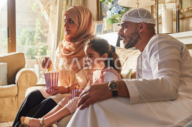 ايماءات الاندماج والتركيز، زوجان عربيان خلجيان اماراتيان يقومان بمشاهدة التلفاز و تناول الفوشار برفقه ابنتهما، أجواء اسرية سعيدة في المنزل، قضاء اوقات عائلية ممتعة، متابعة الافلام والمسلسلات، فلل و شقق سكنية فاخرة في الامارات.