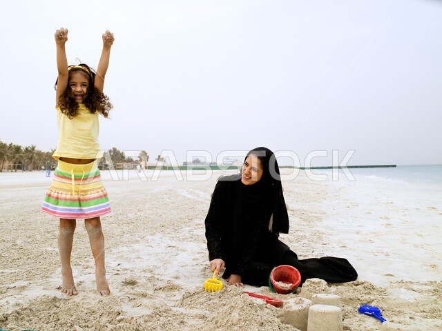 ايماءات وتعابير تدل على السعادة في بناء قلعة الرمال, الجلوس على رمال الشاطئ الناعمة والاستمتاع ببناء المجسمات الرملية, امرأة عربية خليجية سعودية مبتسمة تقضي وقت ممتع برفقة إبنتها على ساحل البحر, قضاء يوم العطلة على البحر، نشاطات ترفيهية عائلية