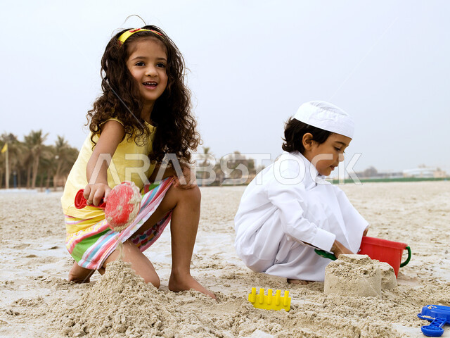 أخ وأخت يلعبان على الشاطئ, فعاليات ترفيهية مسلية للأطفال, قضاء يوم العطلة على البحر, فتاة بإيماءات وجه تدل على السعادة في اللعب برمال البحر الناعمة، طفل عربي خليجي إماراتي يرتدي الثوب السعودي التقليدي يقوم  في بناء المجسمات الرملية