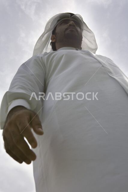 Standing upright and manly, a picture from below of an Emirati Arab Gulf nomad wearing a kandora and a white shemagh standing confidently, looking at something outside under the sun in broad daylight, a view of the blue sky
