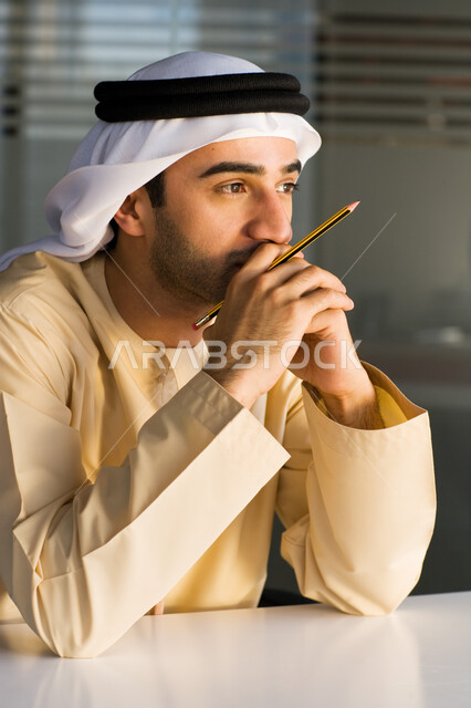 الرؤية المستقبلية والتركيز على ايجاد الحلول، صورة مقربة لرجل عربي خليجي إماراتي يرتدي الغترة والعقال ويجلس أمام المكتب بإيماءات الحيرة والقلق من شيء ما، شرود الذهن والتفكير الزائد في مشكلات العمل، التمعن والتأمل والتخطيط للمستقبل