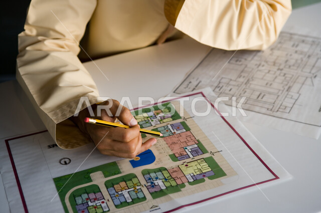 دراسة وتحليل الخرائط الهندسية، صورة مقرة ليد رجل أعمال إماراتي عربي خليجي يمسك القلم ويقوم بتسجيل البيانات والمعلومات اللازمة على إحدى المشاريع العقارية، العمل في الوظائف المكتبية في الشركات الإماراتية