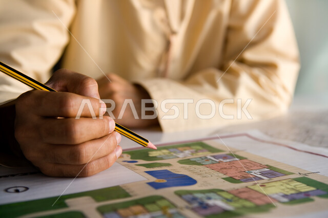 العمل في الوظائف المكتبية في الشركات الإماراتية، صورة مقرة ليد رجل أعمال إماراتي عربي خليجي يمسك القلم ويقوم بتسجيل البيانات والمعلومات اللازمة على إحدى المشاريع العقارية، دراسة وتحليل الخرائط الهندسية