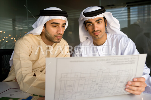 بيئة العمل الإماراتية، التدقيق والإشراف على مخططات مشروع عقاري، اجتماع عمل لرجلان أعمال عربيان خليجيان إماراتيان يرتديان الغترة والكندورة الإماراتية، نقاشات وحوارات حول مخطط خريطة هندسية، مفهوم العمل الجماعي في شركات الإمارات