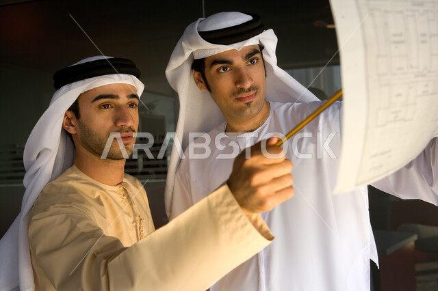 تسجيل البيانات والمعلومات اللازمة على مخطط خريطة هندسية لإحدى المشاريع العقارية، اجتماع عمل لرجلان أعمال عربيان خليجيان إماراتيان يرتديان الغترة والكندورة الإماراتية، مفهوم العمل الجماعي في شركات الإمارات
