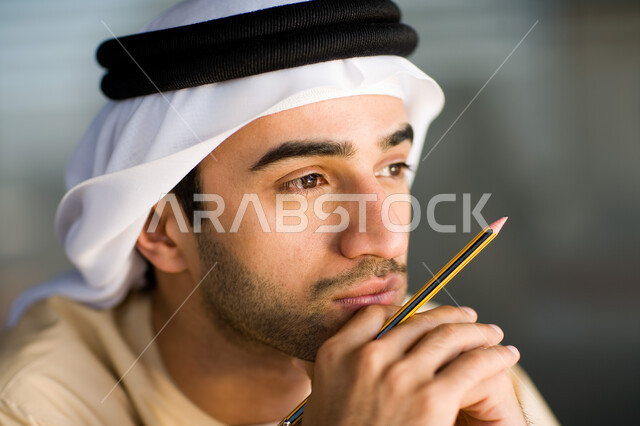 التمعن والتأمل والتخطيط للمستقبل، صورة مقربة لرجل عربي خليجي إماراتي يرتدي الغترة والعقال ويمسك بيده قلم، إيماءات الحيرة والقلق من شيء ما، شرود الذهن والتفكير الزائد في مشكلات العمل، الرؤية المستقبلية والتركيز على ايجاد الحلول
