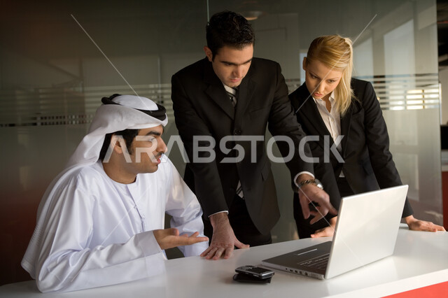 مفهوم الوحدة والعمل الجماعي لتحقيق أهداف الشركة، رجل وسيدة أعمال أجنبيان يستخدمان الحاسوب المحمول في شرح إستراتيجية العمل مع رجل عربي خليجي إماراتي يرتدي العقال والكندورة الإماراتية، تدريب وتطوير الموظفين في الشركات الإماراتية