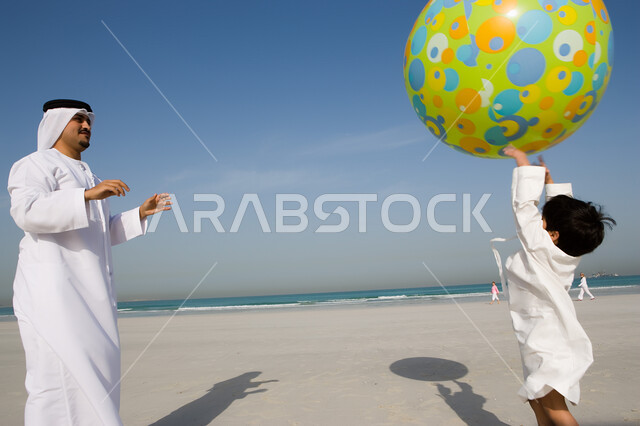 الإستمتاع في قضاء الوقت على شواطئ البحر في الإمارات، أب عربي خليجي إماراتي يرتدي الزي الإماراتي التقليدي ويلعب مع إبنه بالكرة البلاستيكية الكبيرة، مشاركة الأبن في التسلية والترفيه، مفهوم الأبوة وحب الأبناء