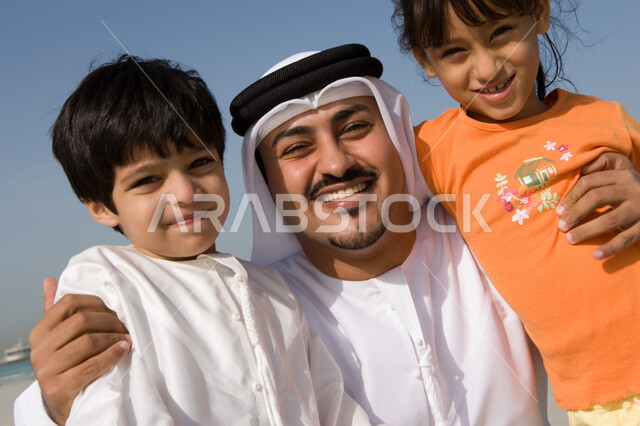 مفهوم الترابط الأسري بين الأب والأبناء، صورة مقربة لرجل عربي خليجي إماراتي يرتدي الزي الإماراتي التقليدي ويقوم بإحتضان أبنائه، الأجواء العائلية المليئة بالحب، إيماءات السعادة والفرح بقضاء الوقت مع الأسرة