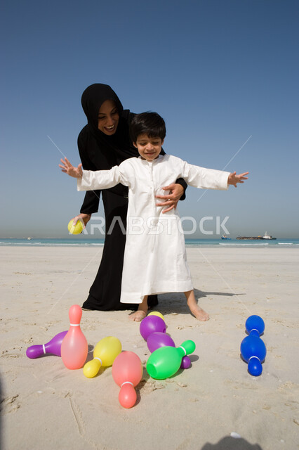 Various recreational activities for children, a veiled Arab Gulf Emirati mother wearing a black abaya accompanied by her child on the soft sand of the beach, gestures of pleasure and happiness by spending time in making the son happy, using plastic bowlin