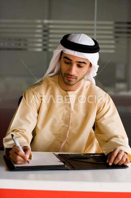 دراسة ومراجعة المشاريع، رجل أعمال عربي خليجي إماراتي يرتدي الغترة والكندورة الإماراتية ويقوم بتدوين وكتابة الملاحظات على الورق، التحقق والتدقيق في شروط عقد عمل جديد، التوقيع على عقود العمل، بيئة العمل في الشركات الإماراتية