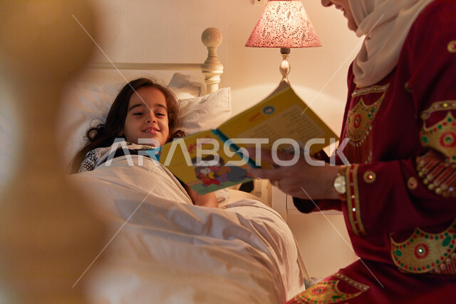 الشعور بالنعاس عند سماع الحكايه، مفهوم السعاده الأسريه وإهتمام الأم بالأبناء، إمرأة عربيه خليجيه إماراتية محجبة ترتدي عباءه مطرزه تقوم بقراءة القصص المسليه لابنتها، الأنشطه الترفيهية قبل النوم، الإستمتاع بالأوقات العائليه في الليل.