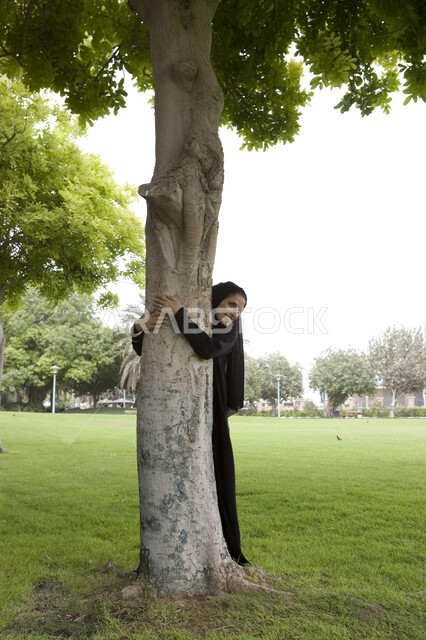 صورة الجسم بالطول الكامل, قضاء أوقات ممتعة في إحدى الحدائق العامة, شابة عربية خليجية إماراتية محجبة تقوم بعناق جذع شجره, اللعب والترفيه في االهواء الطلق, التنزه في الحدائق الخضراء, الاستمتاع في التسلية والترفيه في المنتزهات, اللعب في أوقات الفراغ